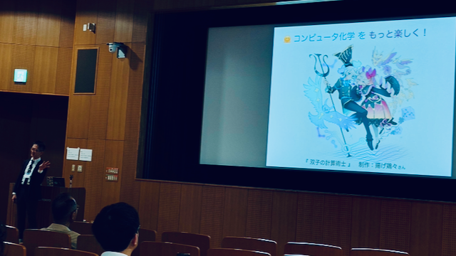 講堂のスクリーンに映したスライド。やまラボのマスコット「双子の計算術士」と「コンピュータ化学 をもっと楽しく！」の一枚